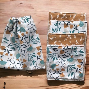 May include: Set of five white cotton face cloths with a floral and bird pattern in shades of green, blue, and yellow. The face cloths are layered on top of each other.