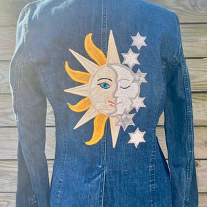 Puede incluir: Chaqueta vaquera azul con un diseño celestial en la espalda. El diseño presenta un sol sonriente, una luna guiñando un ojo y varias estrellas en tonos amarillos, blancos y grises. La chaqueta es de un azul medio.