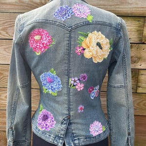Op de afbeelding: Een spijkerjack met geborduurde bloemen in tinten roze, geel, blauw en paars. De bloemen zijn in een patroon op de achterkant van het jack gerangschikt.