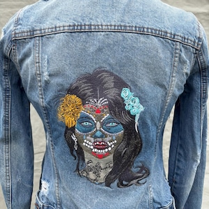 Puede incluir: Chaqueta vaquera azul claro con diseño desgastado. La parte trasera presenta un diseño bordado de calavera de azúcar con detalles en azul, amarillo y rojo. La chaqueta tiene cuello clásico, cierre de botones y mangas largas.