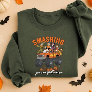May include: A military green crewneck sweatshirt featuring a graphic of Disney characters riding an orange monster truck. The text above reads SMASHING and the text below reads pumpkins in white script. The design includes pumpkins and autumn leaves.