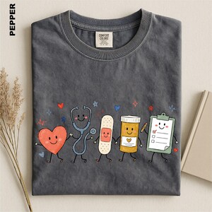 A gray t-shirt with a whimsical graphic design depicting a stethoscope, heart, bandage, pill bottle, clipboard, and a smiling face, all accompanied by colorful stars and hearts.