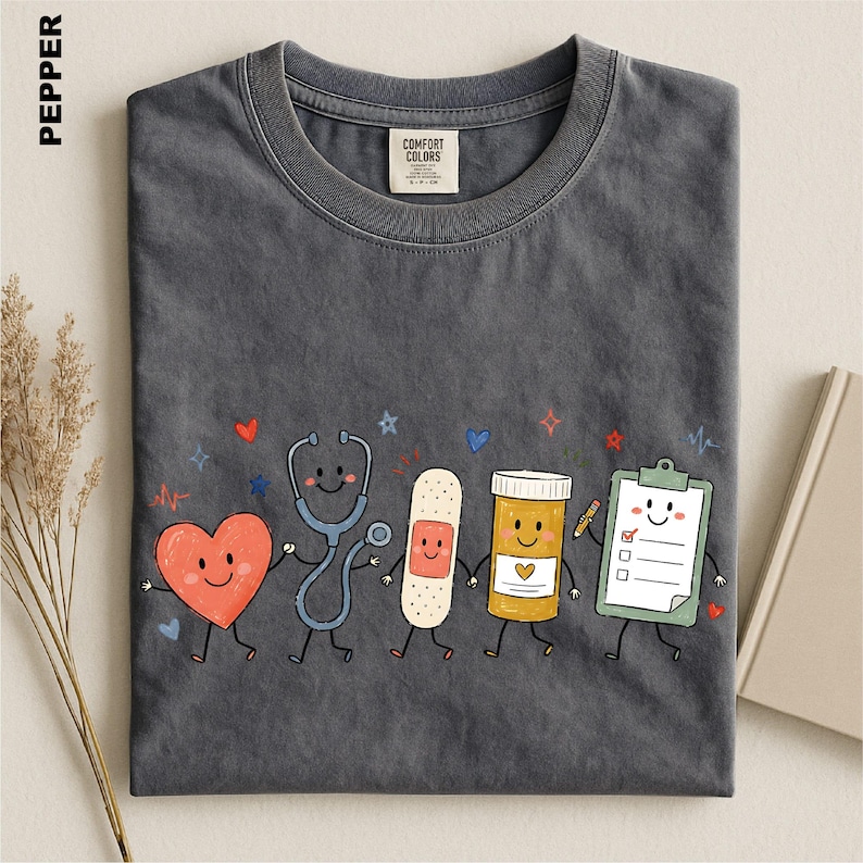 A gray t-shirt with a whimsical graphic design depicting a stethoscope, heart, bandage, pill bottle, clipboard, and a smiling face, all accompanied by colorful stars and hearts.