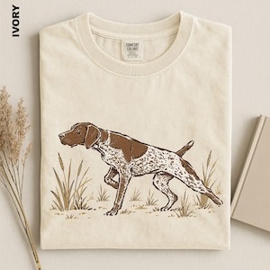 May include: An ivory t-shirt featuring a brown and white graphic illustration of a pointing dog in a field of tall grass. The shirt has a crew neckline and a Comfort Colors brand label on the inside collar. The word IVORY is printed in black.