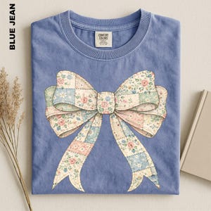 A blue t-shirt with a large bow design on it, placed on a surface alongside some dried flowers and a book.