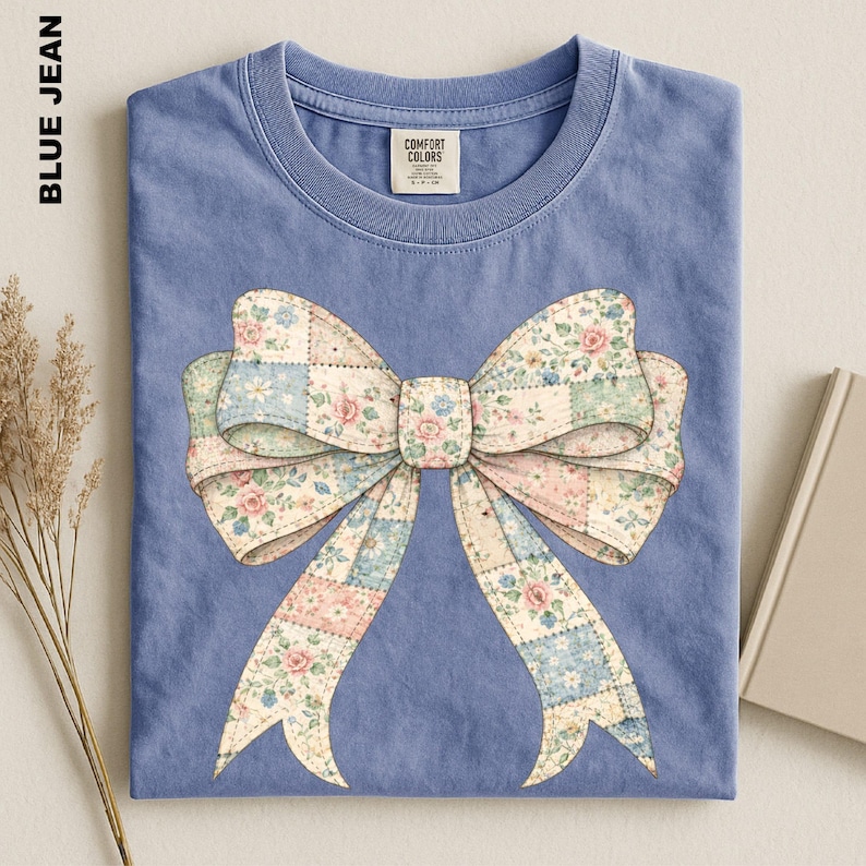 A blue t-shirt with a large bow design on it, placed on a surface alongside some dried flowers and a book.