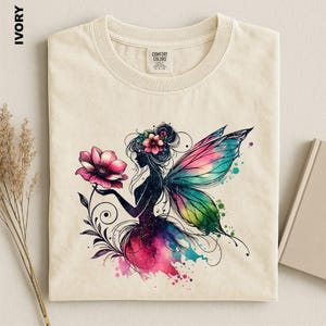 May include: An ivory t-shirt featuring a graphic of a fairy with colorful wings and a flower. The fairy has a dark silhouette with pink and blue watercolor accents. The shirt has a crew neckline and a Comfort Colors brand label at the neck.