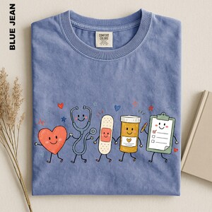 A blue t-shirt with a colorful illustration of cartoon-style medical professionals, including a stethoscope, heart, bandage, pill bottle, clipboard, and a smiling face.