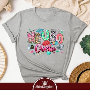 a gray t-shirt with a colorful graphic design that says "Neuro Crew" and includes various medical and healthcare-related icons. The t-shirt is displayed on a white background, surrounded by other items such as a straw hat, a potted plant, and a woven basket.