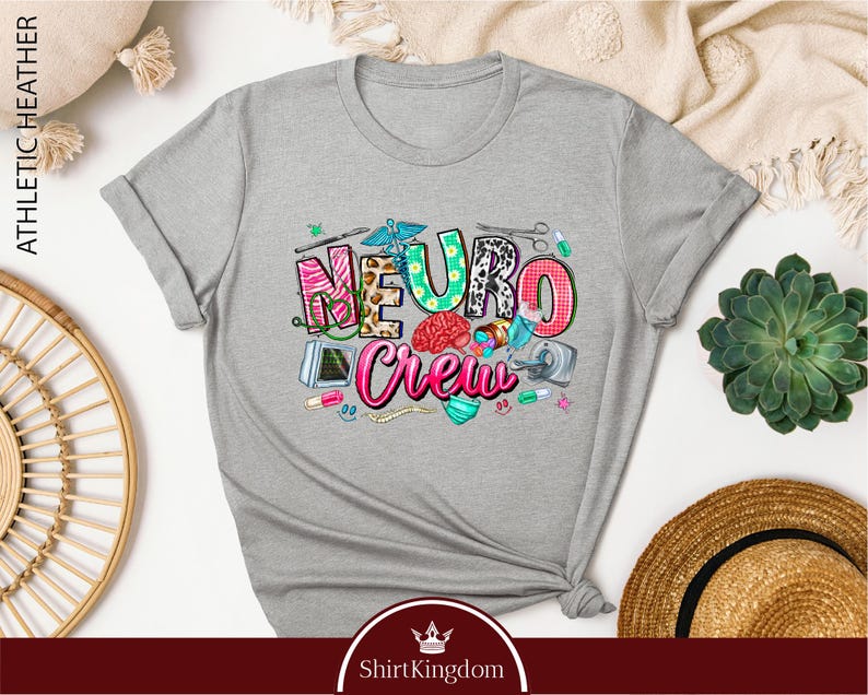 a gray t-shirt with a colorful graphic design that says "Neuro Crew" and includes various medical and healthcare-related icons. The t-shirt is displayed on a white background, surrounded by other items such as a straw hat, a potted plant, and a woven basket.