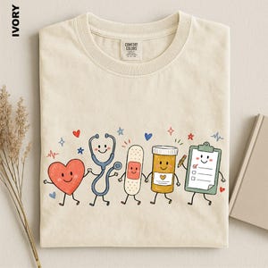 May include: An ivory t-shirt featuring a graphic of five anthropomorphic medical items holding hands: a red heart, a blue stethoscope, a beige bandage, an orange pill bottle, and a green clipboard with a checklist, surrounded by small stars and hearts.