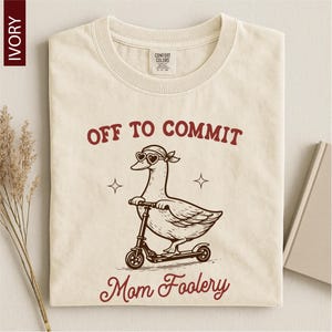 May include: Ivory-colored t-shirt with a graphic of a goose wearing heart-shaped sunglasses and a bandana, riding a scooter. The text "OFF TO COMMIT" and "Mom Foolery" are printed in maroon.