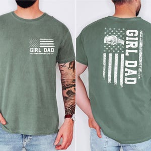 May include: A moss green short-sleeve t-shirt featuring a white distressed American flag graphic with the text GIRL DAD on the front left chest and a larger version of the same graphic with a fist bump illustration on the back.