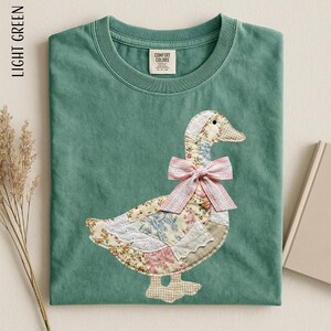May include: Light green t-shirt with a goose applique. The goose is made of floral and patterned fabrics, with a pink gingham bow. The shirt has a crew neck and a Comfort Colors tag.