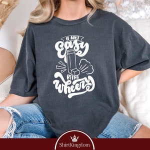 May include: A dark gray t-shirt with white text that reads "It ain't easy being wheezy". The text is stylized with a whimsical font and includes an illustration of an inhaler.