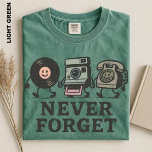May include: A light green t-shirt featuring a graphic of a vinyl record, a vintage camera, and a rotary telephone holding hands. The text NEVER FORGET is printed in black below the illustrations. The shirt has a Comfort Colors brand label.