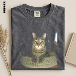 May include: A charcoal gray crew neck t-shirt featuring a graphic print of a tabby cat sitting at a computer keyboard. The shirt has a Comfort Colors brand label at the inner neckline and the word PEPPER printed in the upper left corner.