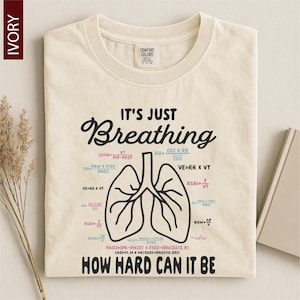 May include: An ivory crew neck t-shirt featuring a black line drawing of human lungs in the center. The text reads IT'S JUST Breathing HOW HARD CAN IT BE, surrounded by various medical respiratory formulas in blue, pink, and black ink.