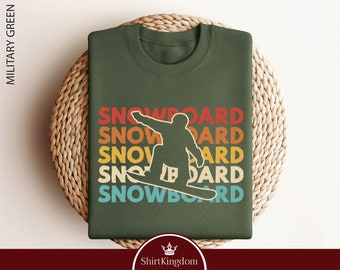 Könnte beinhalten: Ein grünes T-Shirt mit einer Retro-Grafik eines Snowboarders und dem Wort "SNOWBOARD", das in einem Regenbogen-Farbschema wiederholt wird.