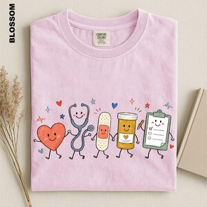 A pink t-shirt with a cartoon illustration of a stethoscope, heart, bandage, pill bottle, clipboard, and other medical-themed characters.