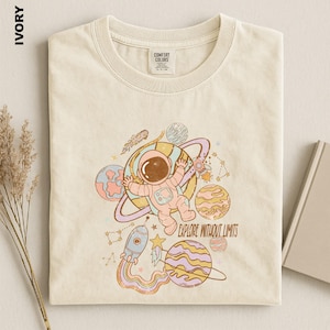 May include: An ivory t-shirt featuring a graphic of an astronaut floating among planets, stars, and a rocket ship. The design includes the text "EXPLORE WITHOUT LIMITS" in a brown font. The shirt has a crew neckline and a Comfort Colors brand label.