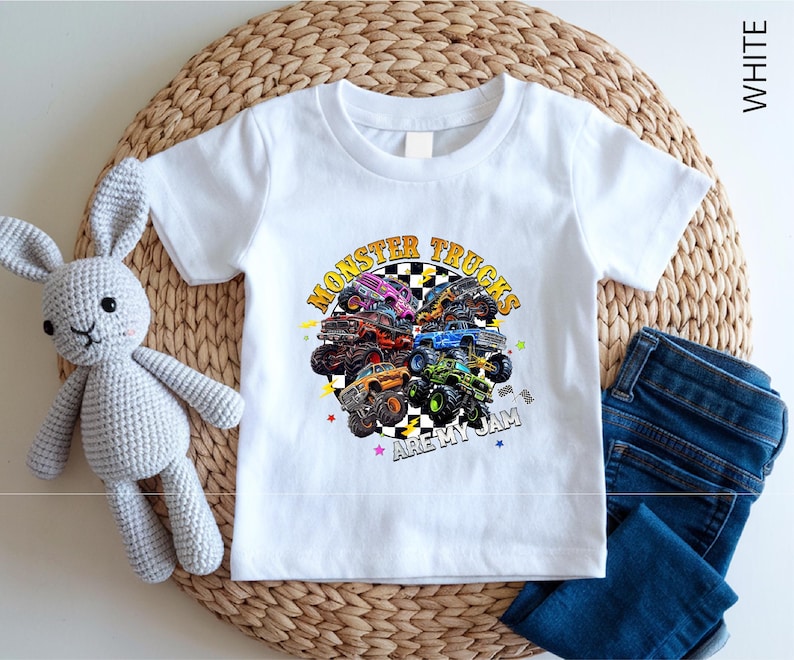 A white t-shirt with a graphic of monster trucks and the text "MONSTER TRUCKS ARE MY JAM" printed on it, along with a crocheted rabbit toy and a pair of blue jeans.