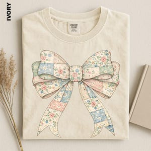 A beige t-shirt with a large floral bow design printed on it, placed on a white surface next to a book and some dried flowers.
