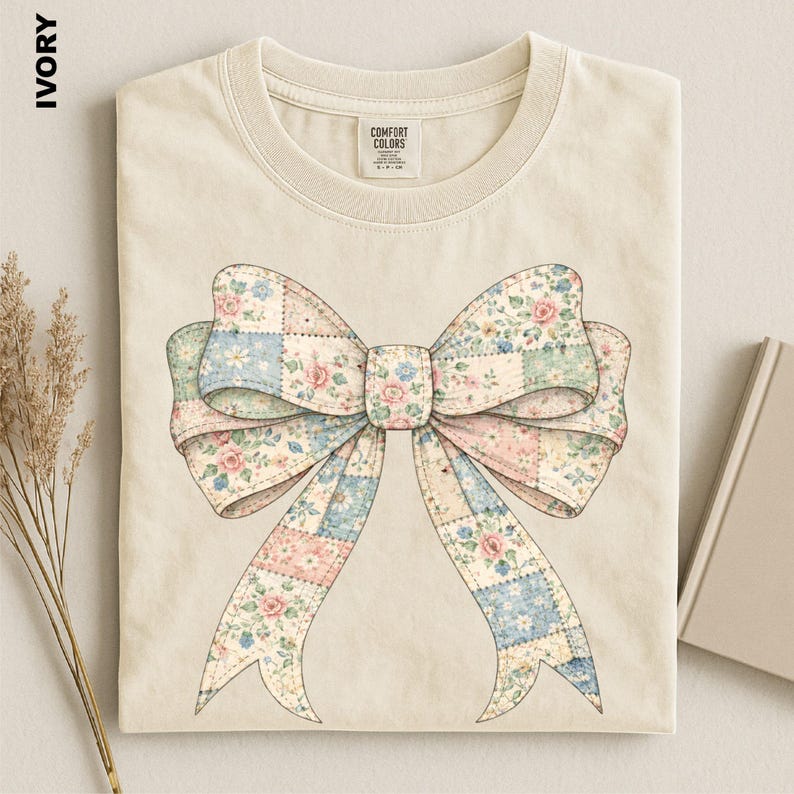A beige t-shirt with a large floral bow design printed on it, placed on a white surface next to a book and some dried flowers.