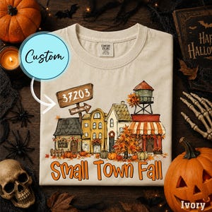 May include: An ivory crew neck t-shirt featuring a graphic of a small town street scene with autumn trees, pumpkins, and a water tower. A wooden signpost displays the text 37203 and Nashville, TN. The text Small Town Fall is printed below.