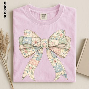 A pink t-shirt with a large bow design printed on it, placed on a surface alongside some dried flowers and a book.
