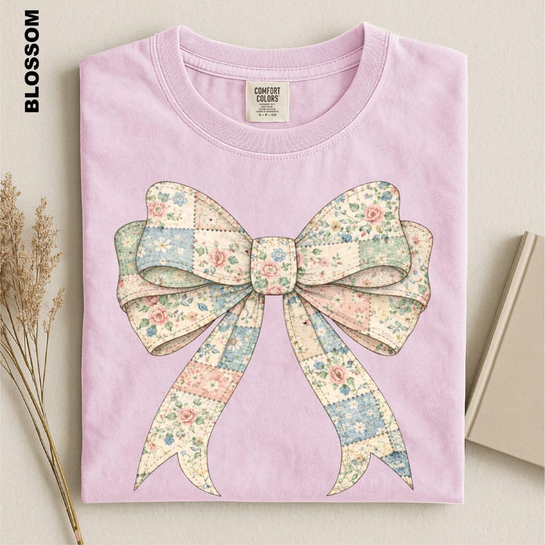 A pink t-shirt with a large bow design printed on it, placed on a surface alongside some dried flowers and a book.