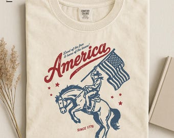 Puede incluir: Camiseta de color marfil con un diseño patriótico. El gráfico presenta la palabra "America" en rojo, un jinete a caballo sosteniendo una bandera estadounidense, y el texto "Land of the free & home of the brave!" y "SINCE 1776".
