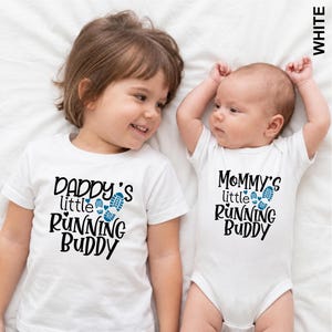 May include: A white short-sleeved t-shirt with black text reading DADDY'S little RUNNING BUDDY and a white baby bodysuit with black text reading Mommy's little RUNNING BUDDY, both featuring blue footprints and small heart graphics.