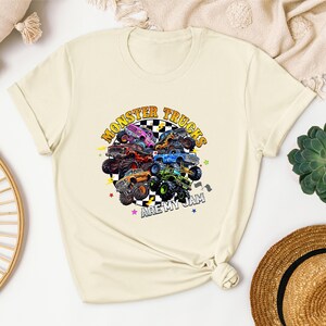 a white t-shirt with a colorful graphic design of monster trucks, along with the text "MONSTER TRUCKS" and "ARE MY JAM" printed on it. The t-shirt is displayed on a white surface, surrounded by various items such as a hat, a woven basket, and a potted plant.