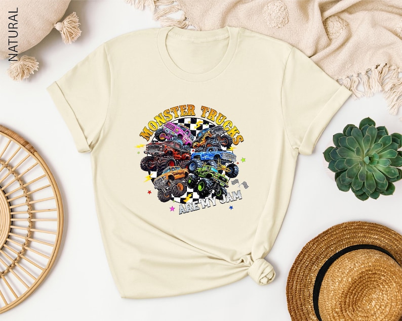a white t-shirt with a colorful graphic design of monster trucks, along with the text "MONSTER TRUCKS" and "ARE MY JAM" printed on it. The t-shirt is displayed on a white surface, surrounded by various items such as a hat, a woven basket, and a potted plant.