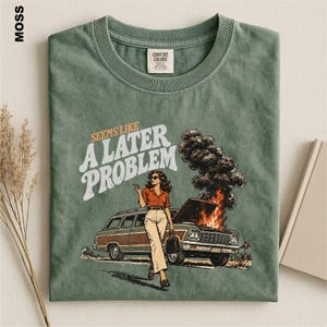 May include: A moss green t-shirt featuring a graphic of a person standing in front of a station wagon with its hood open and smoke billowing out. The text reads "SEEMS LIKE A LATER PROBLEM" in a retro font. The shirt has a crew neck and a label.