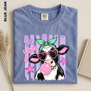 May include: A blue jean colored t-shirt featuring a graphic of a cow wearing a mint green hair bow and pink heart-shaped sunglasses while blowing a pink bubble. The text "be you" is printed in pink next to the cow, surrounded by purple stripes.