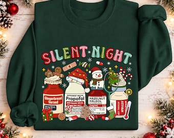 Könnte beinhalten: Ein dunkelgrüner Rundhals-Pullover mit dem Schriftzug "SILENT NIGHT" in bunten Buchstaben. Das Motiv zeigt vier Medikamentenflaschen mit den Aufschriften "Gingerbread Versed", "Peppermint Mocha Propofol", "Hazelnut Haldol" und "Ativan", verziert mit Weihnachtssymbolen wie einem Schneemann, Lebkuchenmann, Zuckerstangen und einem Stethoskop.