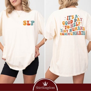May include: A white t-shirt with the text "SLP" in colorful retro font on the front and "It's a good day to help tiny humans communicate" in colorful retro font on the back.