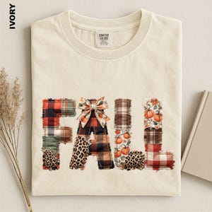 May include: An ivory t-shirt featuring the word FALL in large letters. The letters are composed of various fabric patterns including red and black plaid, brown plaid, leopard print, and a pumpkin print. A decorative bow sits atop the letter A.