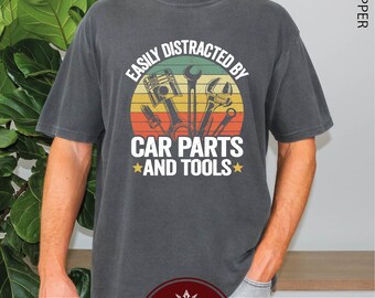 Puede incluir: Una camiseta gris oscuro con un gráfico de estilo vintage que dice "Easily Distracted By Car Parts And Tools". El gráfico presenta una puesta de sol con un motor de automóvil, llaves inglesas y otras herramientas.