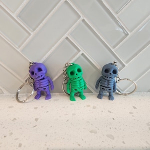 May include: Three colorful skeleton keychains in purple, green, and blue. Each keychain features a small, detailed skeleton figure with a silver keyring attached. The keychains are displayed against a white and gray background.