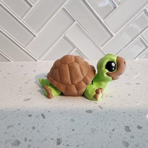 May include: A small, green turtle figurine with brown shell and large, black eyes. The turtle is made of a smooth, painted material and has a friendly expression.
