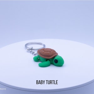 May include: A green and brown turtle keychain with a silver chain. The turtle is small and has a cute expression. The keychain says "BABY TURTLE" on the white surface it is sitting on.
