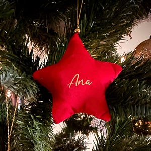 Puede incluir: Un adorno de tela rojo en forma de estrella con el nombre Ana en letras cursivas doradas. El adorno está suspendido por un cordón dorado fino y tiene un acabado suave al tacto con una pequeña cuenta dorada en la parte superior.