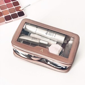 Puede incluir: Un neceser de maquillaje color rosa con la parte superior transparente, que contiene varios productos para el cuidado de la piel. El neceser tiene una cremallera plateada y la letra "A" en la parte delantera. Una paleta de sombras de ojos y un pincel de maquillaje son visibles en el fondo.