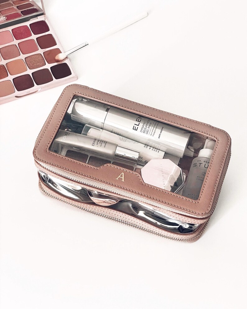 Puede incluir: Un neceser de maquillaje color rosa con la parte superior transparente, que contiene varios productos para el cuidado de la piel. El neceser tiene una cremallera plateada y la letra "A" en la parte delantera. Una paleta de sombras de ojos y un pincel de maquillaje son visibles en el fondo.