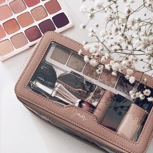 Puede incluir: Una colecci&oacute;n de productos de maquillaje, que incluye paletas de sombras de ojos y un neceser. La bolsa es de color rosa claro con una ventana transparente, que muestra varios art&iacute;culos de belleza. La imagen tambi&eacute;n presenta flores blancas.