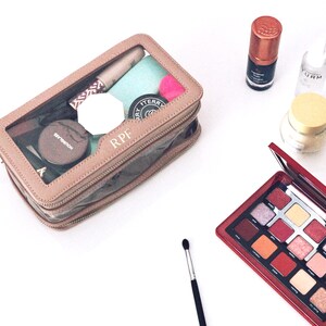 Puede incluir: Un neceser transparente con borde marr&oacute;n, lleno de productos de maquillaje, se encuentra junto a una paleta de sombras de ojos roja. Otros art&iacute;culos incluyen una brocha de maquillaje, un tarro de crema y botellas de productos de belleza.