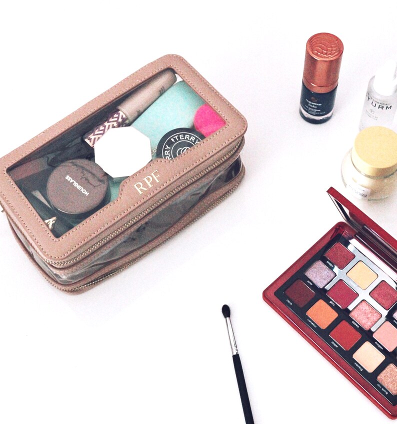 Puede incluir: Un neceser transparente con borde marr&oacute;n, lleno de productos de maquillaje, se encuentra junto a una paleta de sombras de ojos roja. Otros art&iacute;culos incluyen una brocha de maquillaje, un tarro de crema y botellas de productos de belleza.